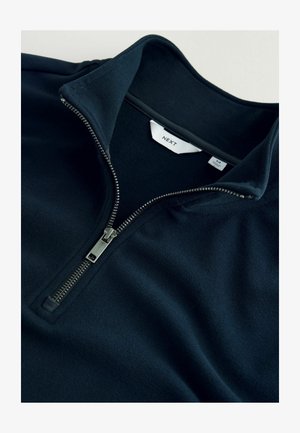 Tume sinine fliisist pullover kõrge kraega, pooleks tõmmatava lukuga ja siledate pindadega. Omab hõbedatooni lukku ja bränditud sildi sees.