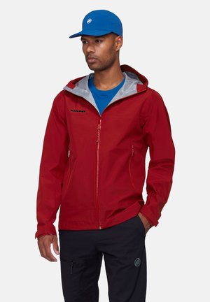 Homme portant une veste à capuche rouge, une chemise bleue, une casquette bleue et un pantalon noir, debout avec une main dans la poche contre un fond uni.