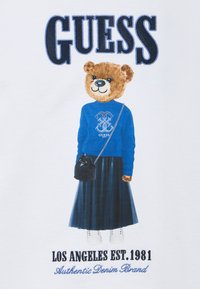 Καρτούν αρκούδα που φοράει μπλε πουλόβερ με το λογότυπο "GUESS", μακριά σκούρα φούστα, λευκά αθλητικά παπούτσια και κρατάει μια μαύρη τσάντα. Λευκό φόντο.