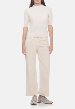Donna con top aderente a maglia costina a maniche corte, pantaloni beige chiaro a gamba larga e sneakers beige, in piedi davanti a uno sfondo semplice.