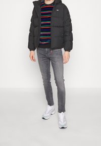 Černá péřová bunda s kapucí na zip. Pod ní vícebarevný pruhovaný svetr a šedé džíny, doplněné bílými teniskami.