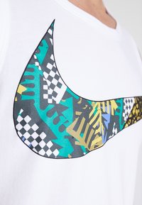 Camiseta blanca con un gran logo de Nike en forma de swoosh multicolor. El logo presenta patrones geométricos en negro, verde azulado, amarillo y azul.
