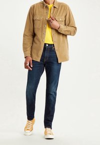 Tan kordfläckad knapprad skjorta med två bröstfickor, gul t-shirt under, mörkblå jeans och gula sneakers med vita sulor.