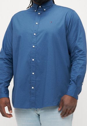 Homme portant une chemise bleue boutonnée à manches longues et un jean clair, montrant le torse et les bras sur fond blanc.