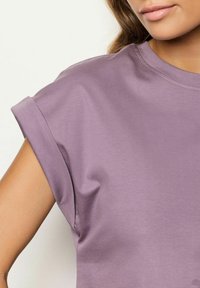 T-shirt viola a maniche corte con scollatura rotonda, maniche arrotolate, texture liscia e leggera drappeggiatura sulla spalla.