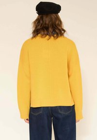 Pull en tricot jaune avec une coupe courte et carrée, et un ourlet côtelé. Présente des épaules tombantes et une couture centrale au dos. Associé à un denim foncé.