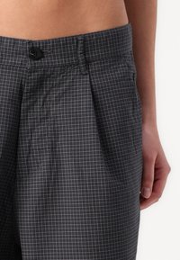 Gray checkered fabric pants with pleats, featuring a black button at the waistband and side pockets. Material has a smooth texture.