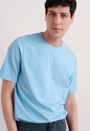 T-shirt bleu clair à manches courtes fait de coton doux. Présente un col rond et une coupe décontractée. Porté avec un pantalon noir à cordon.