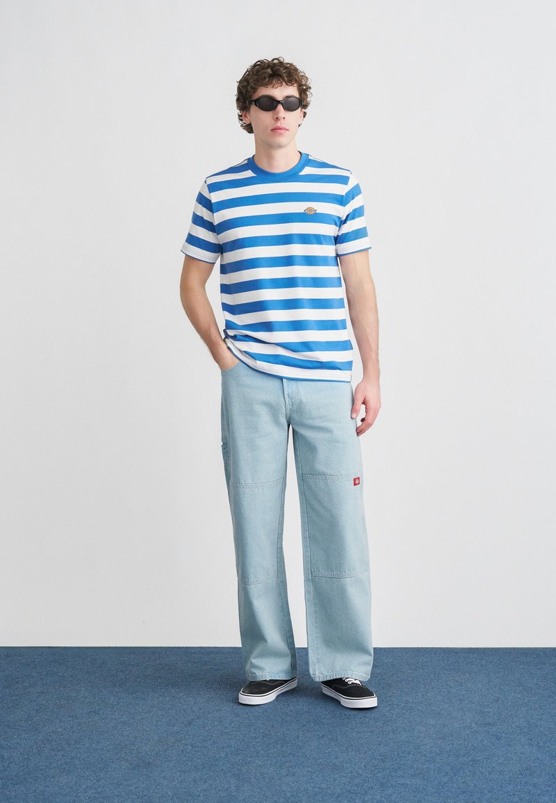 Gestreiftes blau-weißes T-Shirt in entspannter Passform, kombiniert mit hellblauen, weit geschnittenen Hosen. Schwarze Sonnenbrille sowie schwarze und weiße Sneaker vervollständigen das Outfit.