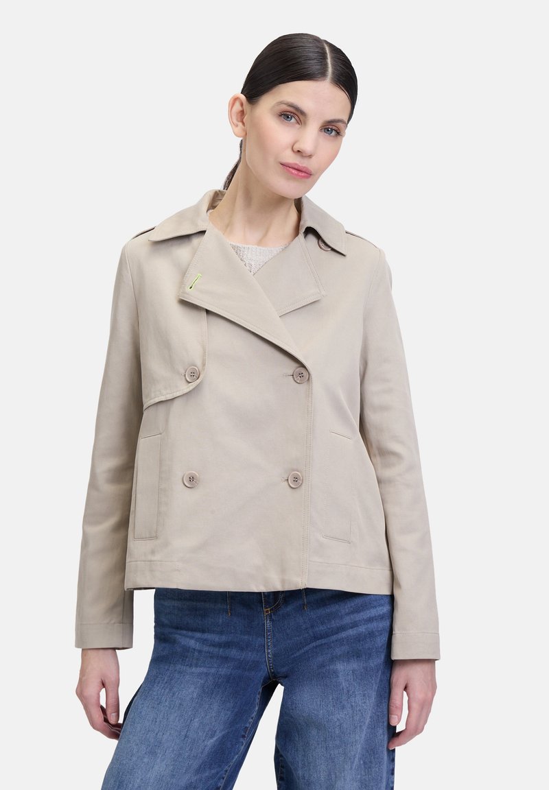 Beige dubbele jas met brede revers, voorzakken en knoopdetails. Gemaakt van een zachte stof. Gecombineerd met blauwe spijkerbroek.