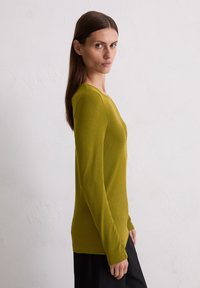 T-shirt à manches longues de couleur vert olive, texture côtelée, col rond, coupe ajustée. Associé à un bas noir, modèle présenté de profil.