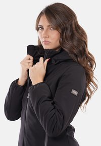 Giacca nera isolata con colletto alto, zip frontale e polsini aderenti. Presenta una patch con il logo sulla manica sinistra. Tessuto liscio.