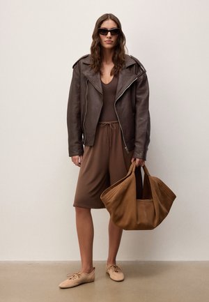 Žena v nadměrné tmavě hnědé kožené bundě, hnědých šatech, béžových botách a slunečních brýlích, držící velkou světle hnědou semišovou tašku, stojící u jednobarevné stěny.