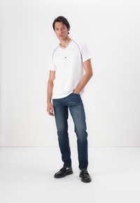 Witte shirt met korte mouwen en een V-hals, met blauwe accenten, gecombineerd met blauwe jeans en zwarte schoenen. Model staat met de handen in de zakken.