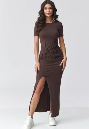 Kobieta ubrana w dopasowaną brązową sukienkę z krótkimi rękawami, z przodem zawiązanym na supeł, bocznym rozcięciem i białymi trampkami, stojąca na jednolitym jasnym tle.