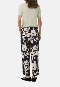 Lichtgroen kortmouwtop; zwarte en crèmekleurige wijde broek met bloemmotief; elastische tailleband; zwarte schoenen. Achteraanzicht.