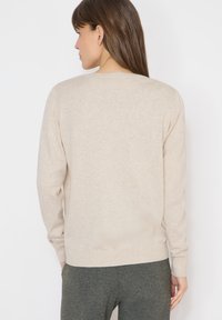 Femme aux longs cheveux bruns portant un pull beige à manches longues et un pantalon gris foncé, debout de dos sur un fond blanc.
