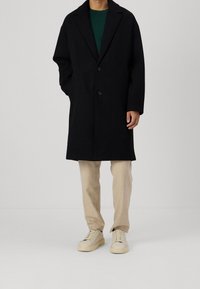 Schwarzer Mantel aus Wollmischung mit Reverskragen, Zwei-Knopf-Verschluss und Seitentaschen, kombiniert mit beigen Hosen und hellen Sneakers.