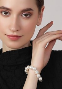 White pearl bracelet with rounded beads, silver accent, worn on a hand against a textured black top. Smooth surface and shiny finish.