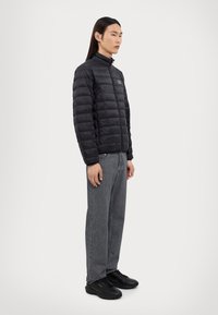 Doudoune noire avec un col haut et un design matelassé horizontal. Portée avec un jean gris et des baskets noires, arborant un logo simple.