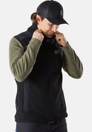 Black fleece vest with textured fabric, featuring a front zipper, side pockets, and a dark green long-sleeve shirt underneath. Black cap worn.