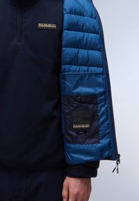 Granatowy polar z rękawami w niebieskie pikowanie. Posiada kieszeń na zamek na rękawie oraz naszywkę z logo na klatce piersiowej. Miękka tekstura na całej powierzchni.