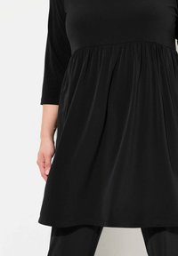 Musta mekko, joka on valmistettu sileästä kankaasta. Mekossa on rento malli, kolmanneksen pituiset hihat ja rypytetty vyötärö. Mallin käsi näkyy.