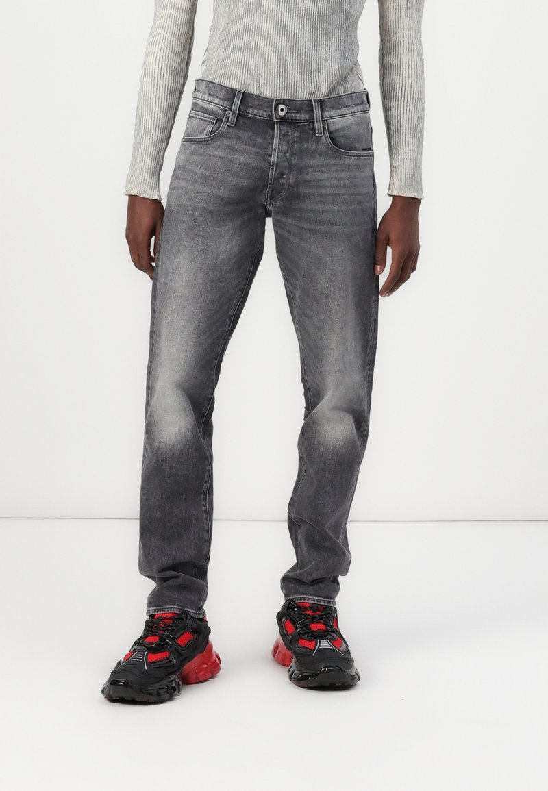Jeans de denim gris con un corte recto, que presentan un ligero desgaste, cinco bolsillos y un cierre de cremallera. Combinados con zapatillas deportivas negras y rojas.