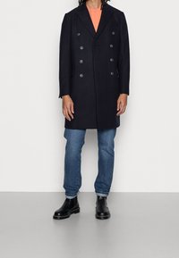 Abrigo azul marino cruzado con seis botones, solapas en pico, llevado sobre una camisa melocotón y jeans azules, combinado con botas negras hasta el tobillo.