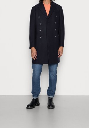 Cappotto navy doppiopetto con sei bottoni, rever a punta, indossato su una camicia pesco e jeans blu, abbinato a stivali neri alla caviglia.