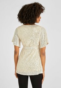 Beiger Top mit Pailletten in lockerer Passform, kurzen Rüschensleeves, tailliertem Gürtel und glatter Textur. Schwarze Hosen ergänzen das Outfit.