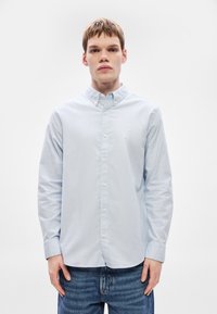 Gaiši zilas krāsas krekli ar garām piedurknēm un pogām, ar apkakli, gludu kokvilnas tekstūru un mazu logo uz krūšu zonas.