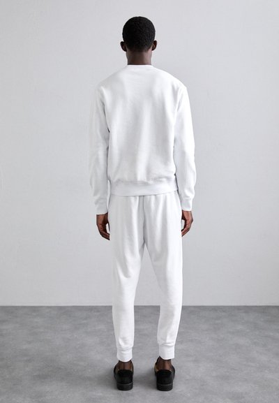 White sweatshirt and joggers, cotton material, ribbed cuffs and waistband, relaxed fit, worn with black footwear, viewed from the back.