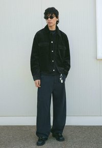 Jachetă din corduroy negru cu două buzunare pe piept, stilizată cu pantaloni largi de culoare închisă și pantofi negri lucioși, asortată cu ochelari de soare ovali.