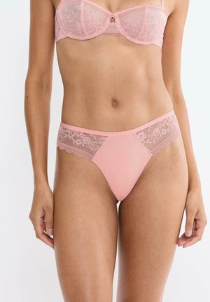Donna con reggiseno di pizzo rosa e mutandine abbinate, in piedi contro uno sfondo bianco semplice, torso e parte superiore delle gambe visibili.