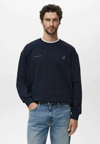 Man met krullend haar in een marineblauwe sweatshirt met kleine tekst en logo op de borst, gecombineerd met lichtblauwe jeans, staand tegen een effen achtergrond.