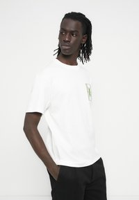 Λευκό βαμβακερό t-shirt με άνετη εφαρμογή, που διαθέτει πράσινο γεωμετρικό λογότυπο στην περιοχή της τσέπης στο στήθος. Κοντά μανίκια, στρογγυλή λαιμόκοψη.