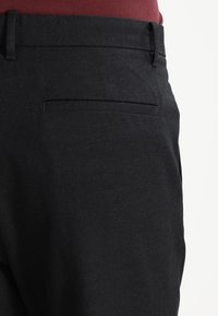 Mustat suorat housut, joissa on sileä kangas ja kapea istuvuus. Takana on yksi tasku, ja vyötärönauha on tummasta, yksivärisestä kankaasta.