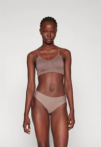 Καφέ κορμάκι με λεπτές τιράντες και χωρίς ραφές, συνδυασμένο με αντίστοιχα γυναικεία σλιπ χαμηλής μέσης. Μαλακό, ελαστικό ύφασμα, με ελάχιστες λεπτομέρειες.