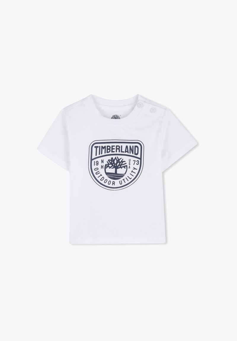 T-shirt blanc à manches courtes en coton, avec un logo Timberland bleu marine accompagné des inscriptions "OUTDOOR UTILITY" et de l'année de création. Deux boutons-pression sur l'épaule.