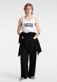 Top sin mangas blanco con el logo de "VANS", pantalones anchos negros y una sudadera negra atada a la cintura. Zapatillas de slip-on de cuadros en blanco y negro.