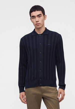 Junger Mann trägt eine marineblaue, gerippte Strickjacke mit Knöpfen und Kragen und beige Hose, steht vor einem schlichten hellen Hintergrund.