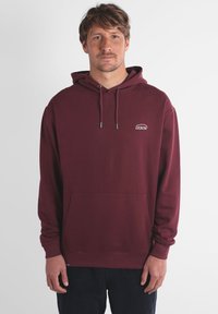 Hoodie bordeaux en tissu doux, doté d'une poche avant, de cordons de serrage et d'un petit logo blanc sur la poitrine. Manches longues.