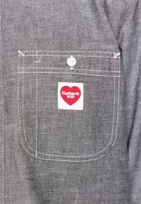 Grijze stoffen zak met witte stiksels en knoop, met een rood hartvormig label waarop "Carhartt WIP" staat, vastgenaaid aan de voorkant van de zak.