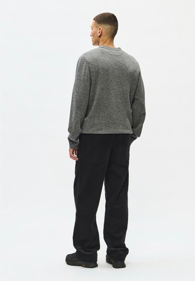 Jeune homme aux cheveux courts portant un pull gris, un pantalon noir et des chaussures noires, debout de dos sur un fond blanc uni.