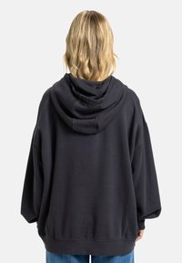 Overgrote zwarte hoodie met een zachte textuur, een trekkoordhoed en geribbelde manchetten, gecombineerd met lichtblauwe jeans. View vanaf de achterkant.