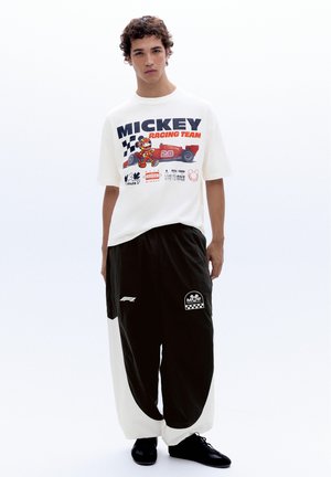 Jeune homme portant un t-shirt blanc Mickey Racing Team et un pantalon large noir et blanc, debout devant un fond blanc uni.