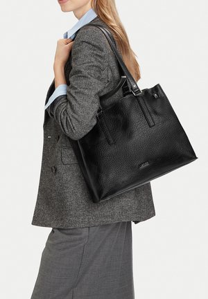 Donna con blazer grigio e pantaloni che porta una grande borsa tote in pelle nera con texture sulla spalla.