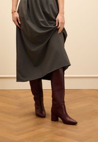 Knielange burgunderrote Lederstiefel mit blockiertem Absatz, ausgestattet mit einer spitzen Zehe und glatter Textur, kombiniert mit einem grauen Stoffrock.