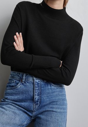Person med armene krydsede iført en sort langærmet trøje og højtaljede blå jeans mod en ensartet lys baggrund.